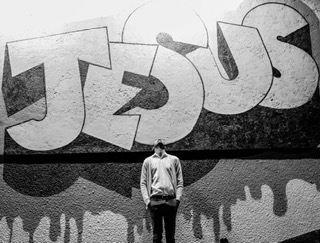 A man stands in front of a high wall. Behind him graffiti with block letters spell 'Jesus'.