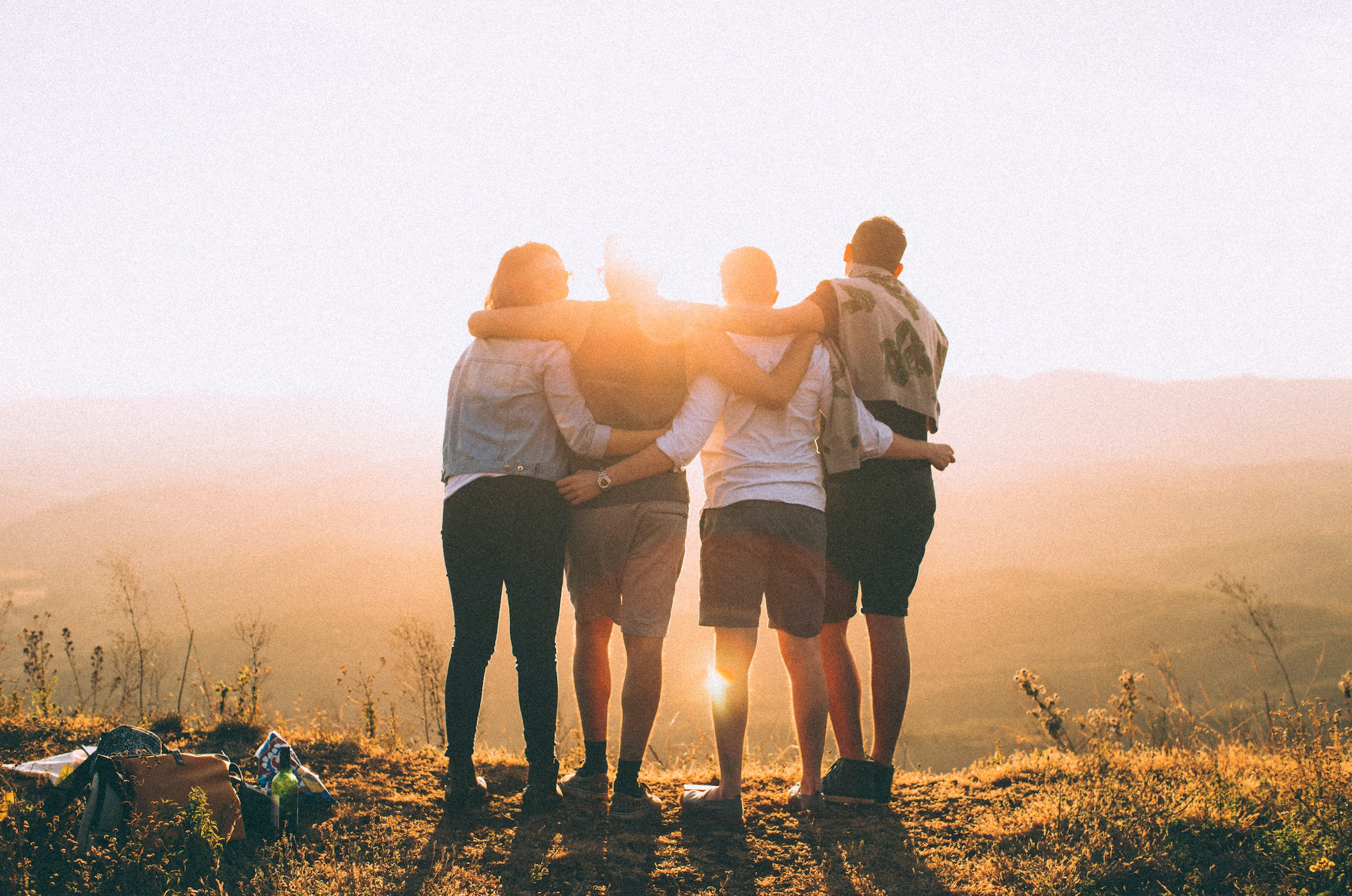Four people, holding each other, with backs to the camera looking towards a view. The sun is behind them.