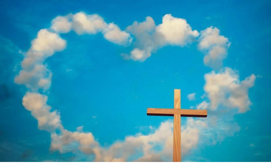 A blue sky with a heart made out of clouds. In the foreground is a wooden cross.