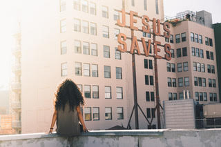 A girl sat on the edge of a building with the words 'Jesus Saves' on a nearby building