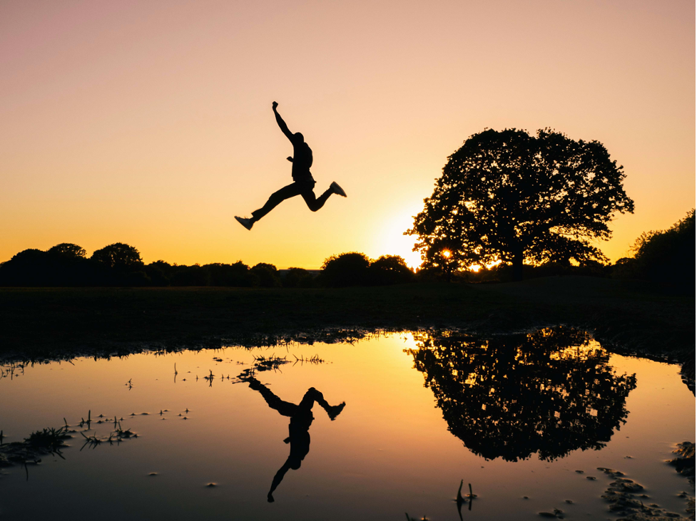 A silhouette of a man in mid air jumping over a large pond
