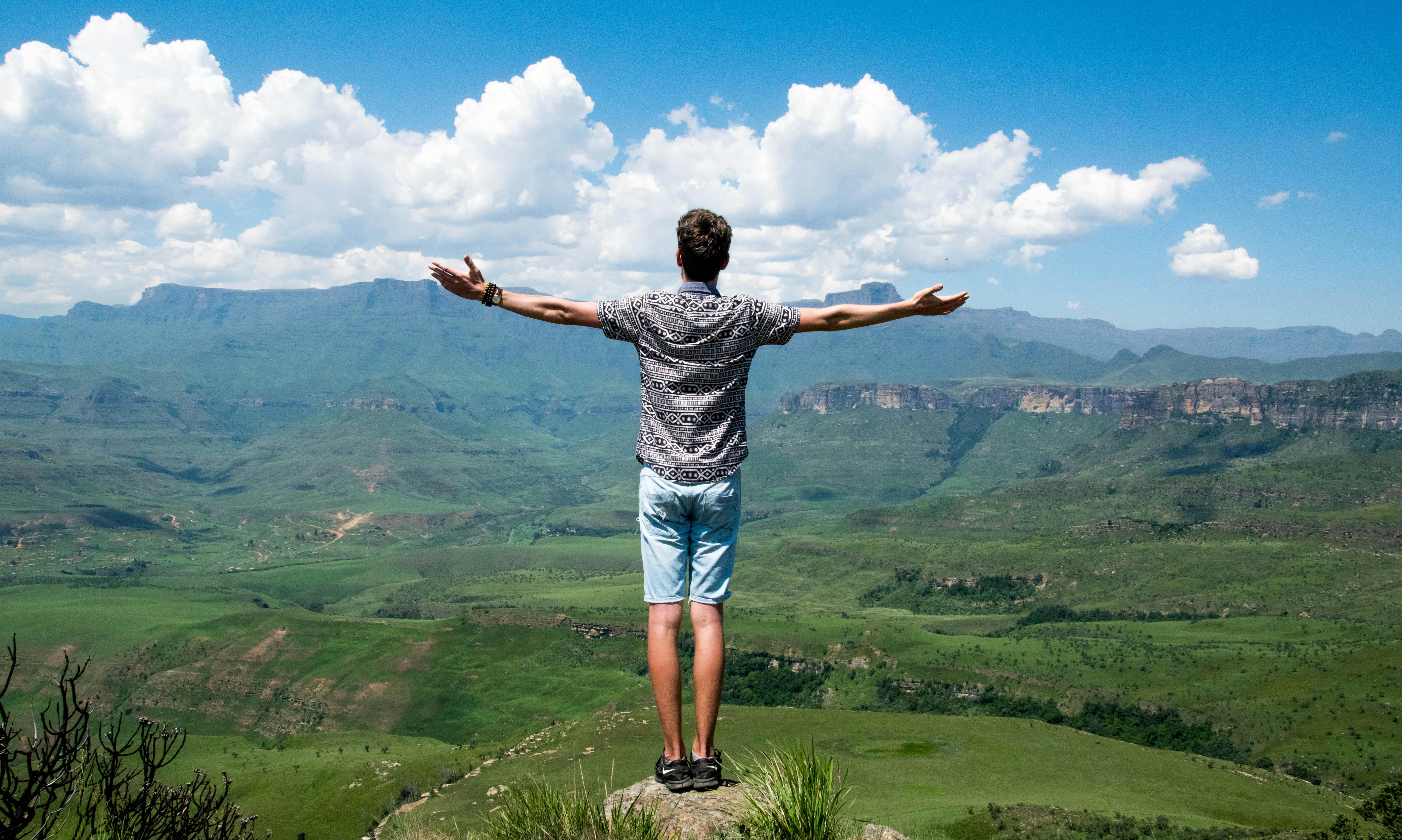Man with back to the camera stood with arms outstretched. The man is up high and there is a view of the countryside and mountains in the distance.