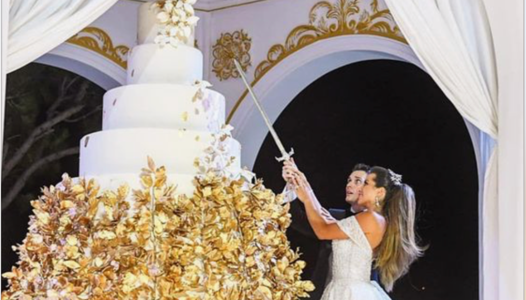 A giant wedding cake. A bride and groom have a sword knife and are holding it up high to try to cut the cake